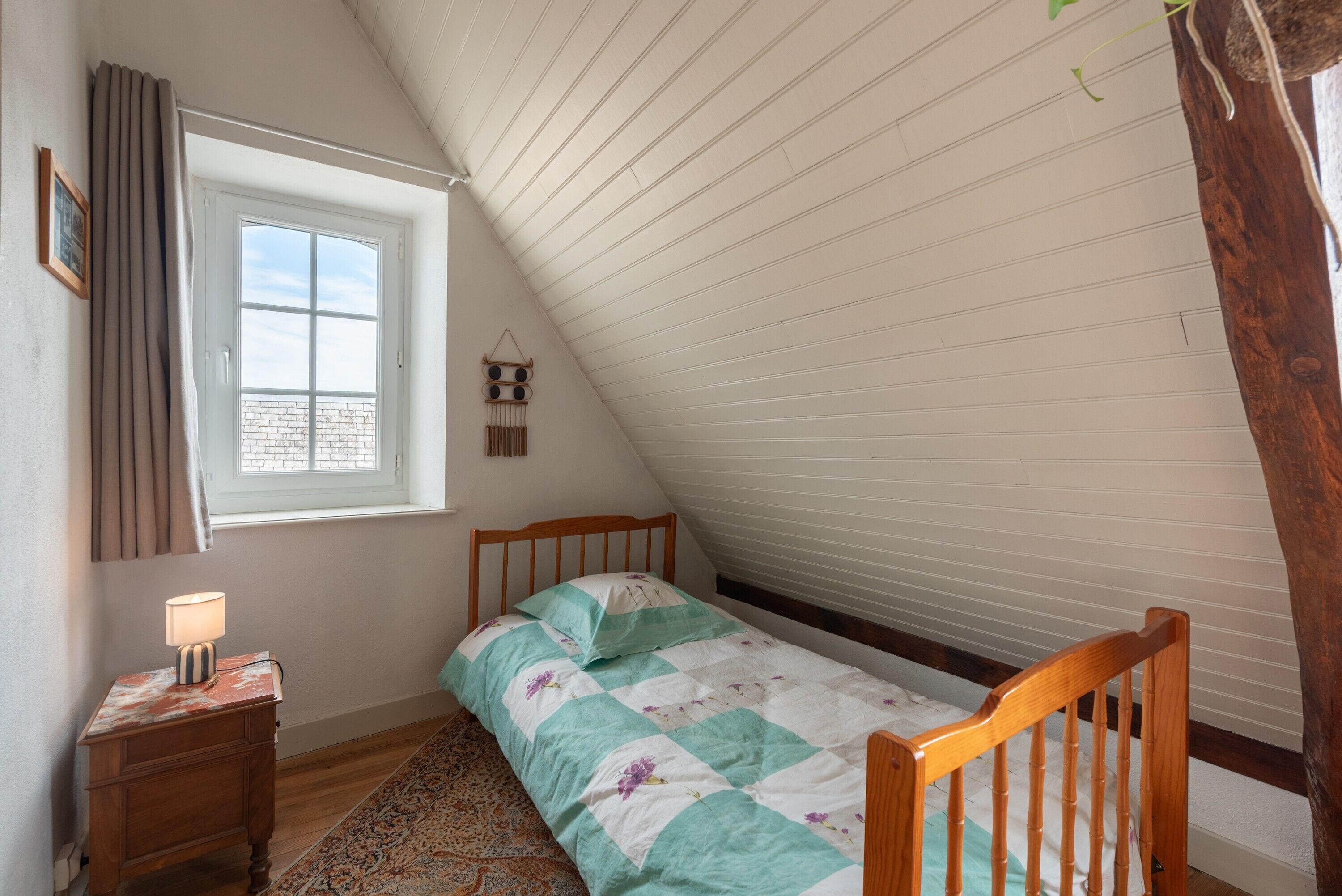 A bedroom with a sloped ceiling, a bed with a quilt, a bedside table with a lamp, a window with curtains, and a wall-mounted shelf.