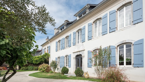 Un bâtiment à deux étages avec une allée pavée, un petit jardin agrémenté d’arbustes et d’un arbre, et des volets bleus aux fenêtres.