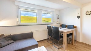 A room with a sofa, a dining table with chairs, a window with a view of a field, a radiator, and a clock on the wall. - Apartment 'Pusteblume' with shared terrace and Wi-Fi (Schmallenberg)
