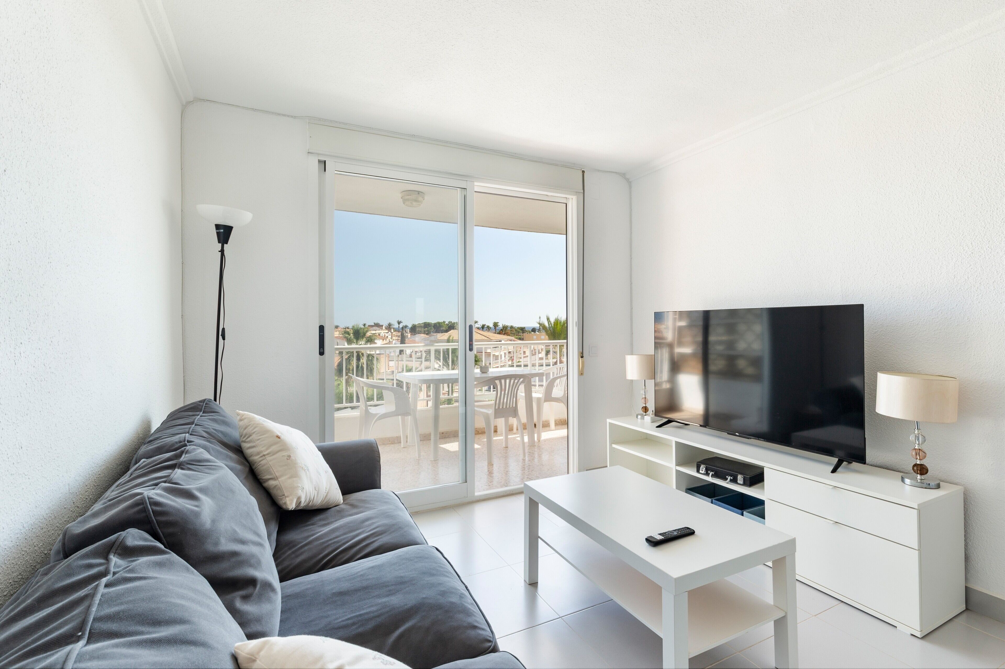 Un salon moderne avec un canapé gris, une table basse blanche, un téléviseur posé sur un meuble blanc et un balcon aménagé avec du mobilier d’extérieur.