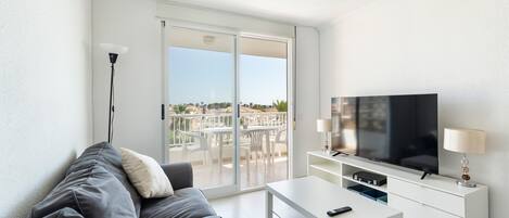 Un salon moderne avec un canapé gris, une table basse blanche, un téléviseur posé sur un meuble blanc et un balcon aménagé avec du mobilier d’extérieur.