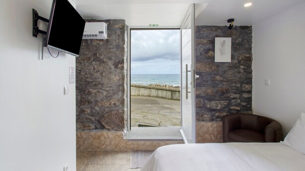 A bedroom with a stone wall, a bed, a TV, and a view of the sea through an open door.