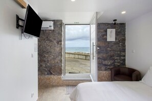 A bedroom with a stone wall, a bed, a TV, and a view of the sea through an open door.