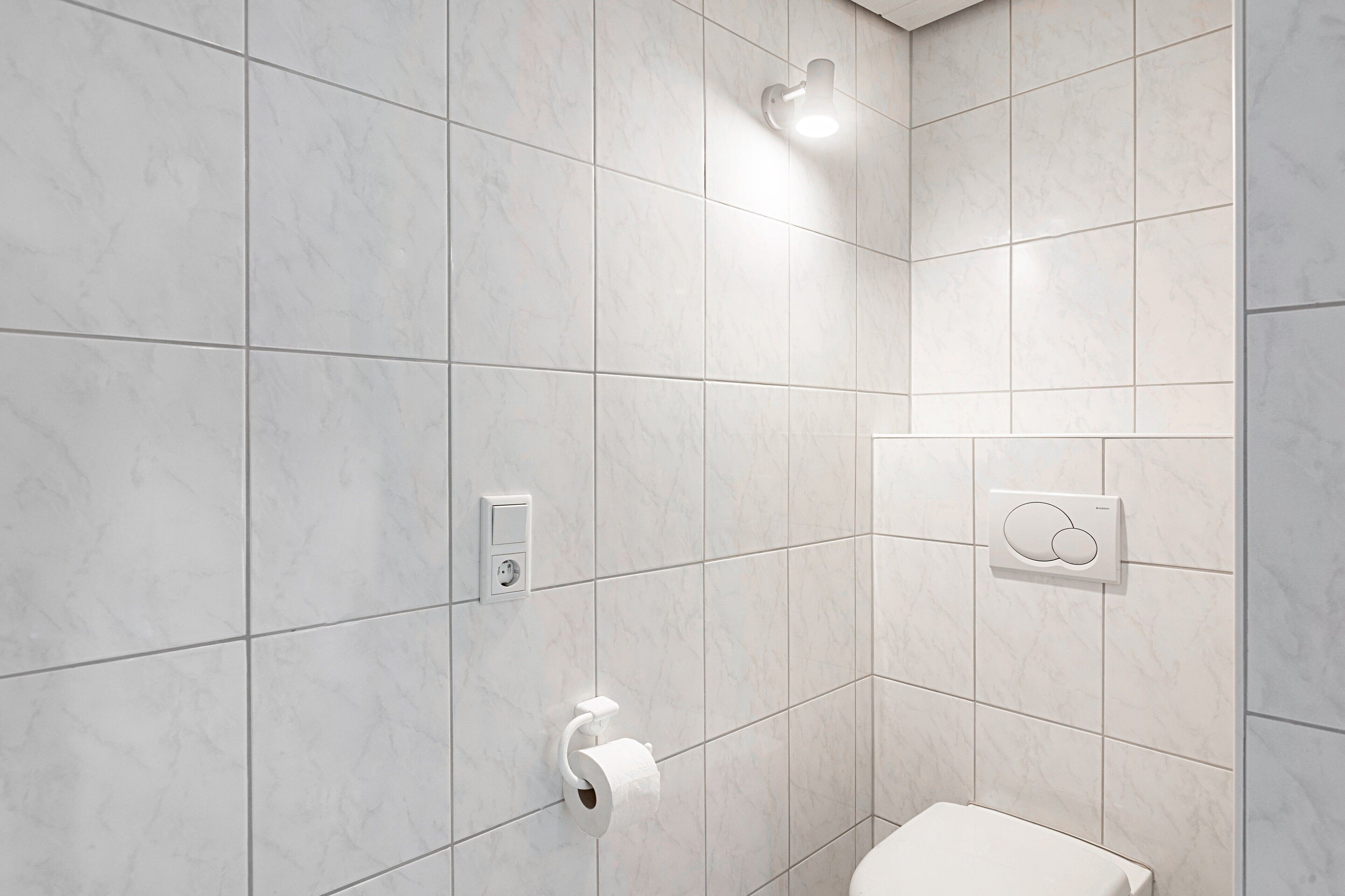 A bathroom with white marble tiles, a toilet, a paper towel dispenser, and a wall-mounted light fixture.