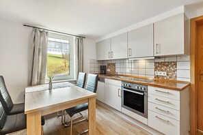 A modern kitchen with white cabinets, a wooden countertop, a dining table with chairs, and a view of a green landscape through a window.