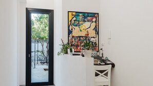 Een hal met een deur naar een buitenruimte, een wit receptiebalie en een kleurrijk abstract schilderij aan de muur.
