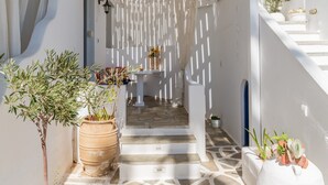 Un étroit sentier mène à une pièce blanche dont le sol est recouvert de carreaux, où se trouvent une petite table avec un vase et des plantes en pot.