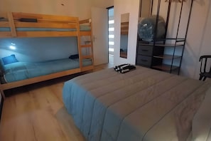 Un lit superposé avec un lit inférieur, un lit simple avec une couvre-lit bleue, une table de chevet avec une lampe et une armoire avec un miroir et des étagères.