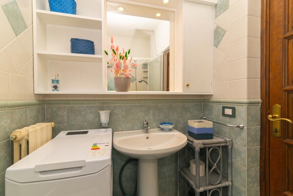 Un baño con un lavamanos blanco, un espejo encima, un perchero, un radiador y una pequeña estantería con una cesta azul y un florero con flores.