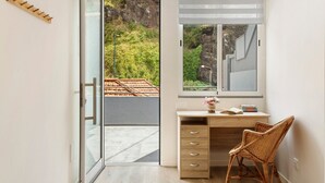 Habitación con puerta de vidrio corredera que da a un balcón, un escritorio con cajones, una silla de mimbre y una ventana con vistas a zonas verdes y un edificio.