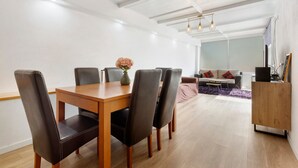 Un área de comedor con una mesa y sillas de madera, un jarrón con flores y un sofá con cojines al fondo.