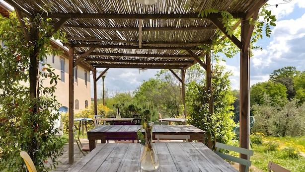 Ein Holztisch mit einer Vase, einer Pergola und Blick auf Grünpflanzen sowie ein Gebäude im Hintergrund.