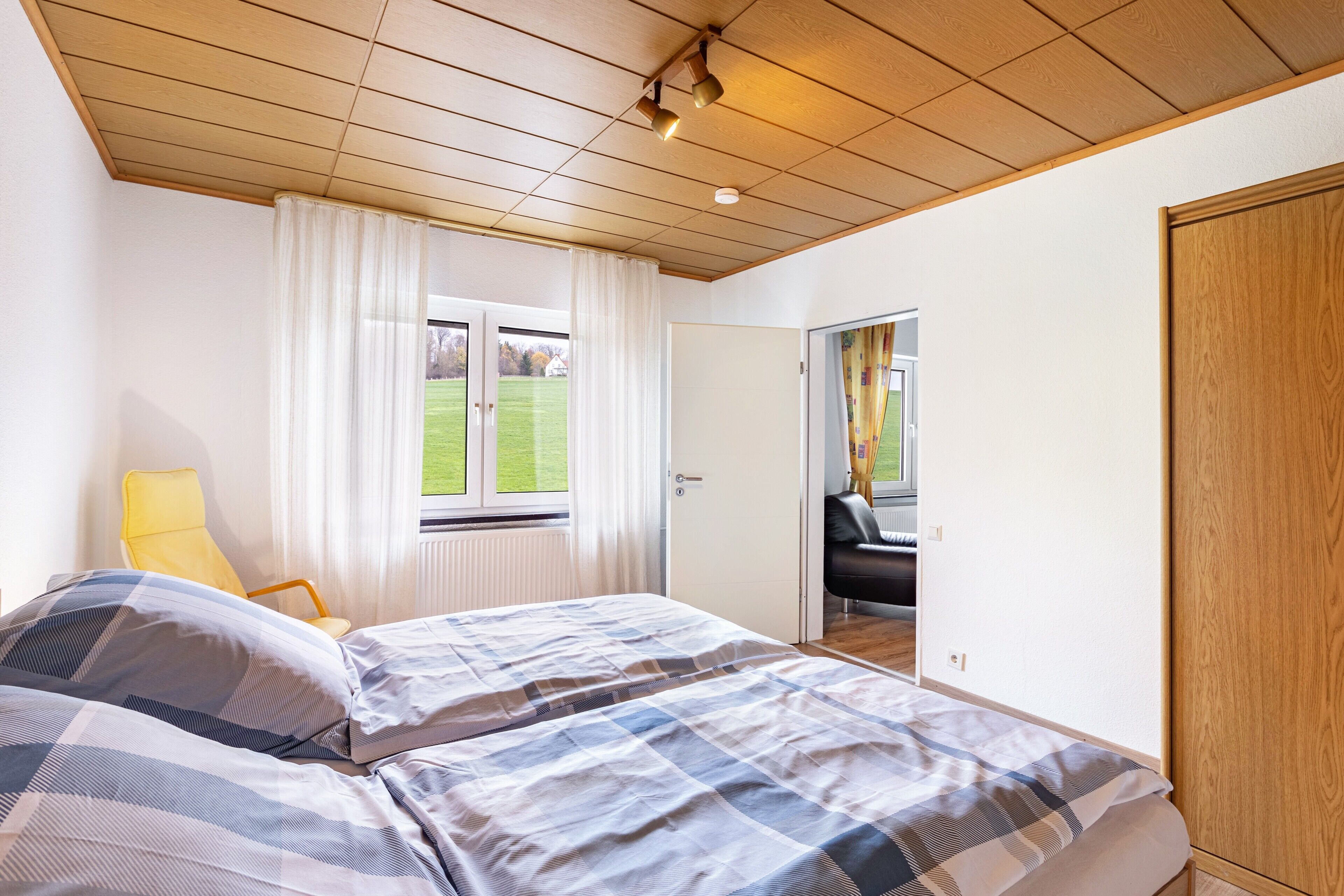 Un dormitorio con una cama, una ventana con cortinas, una puerta que conduce a otra habitación y un armario de madera.
