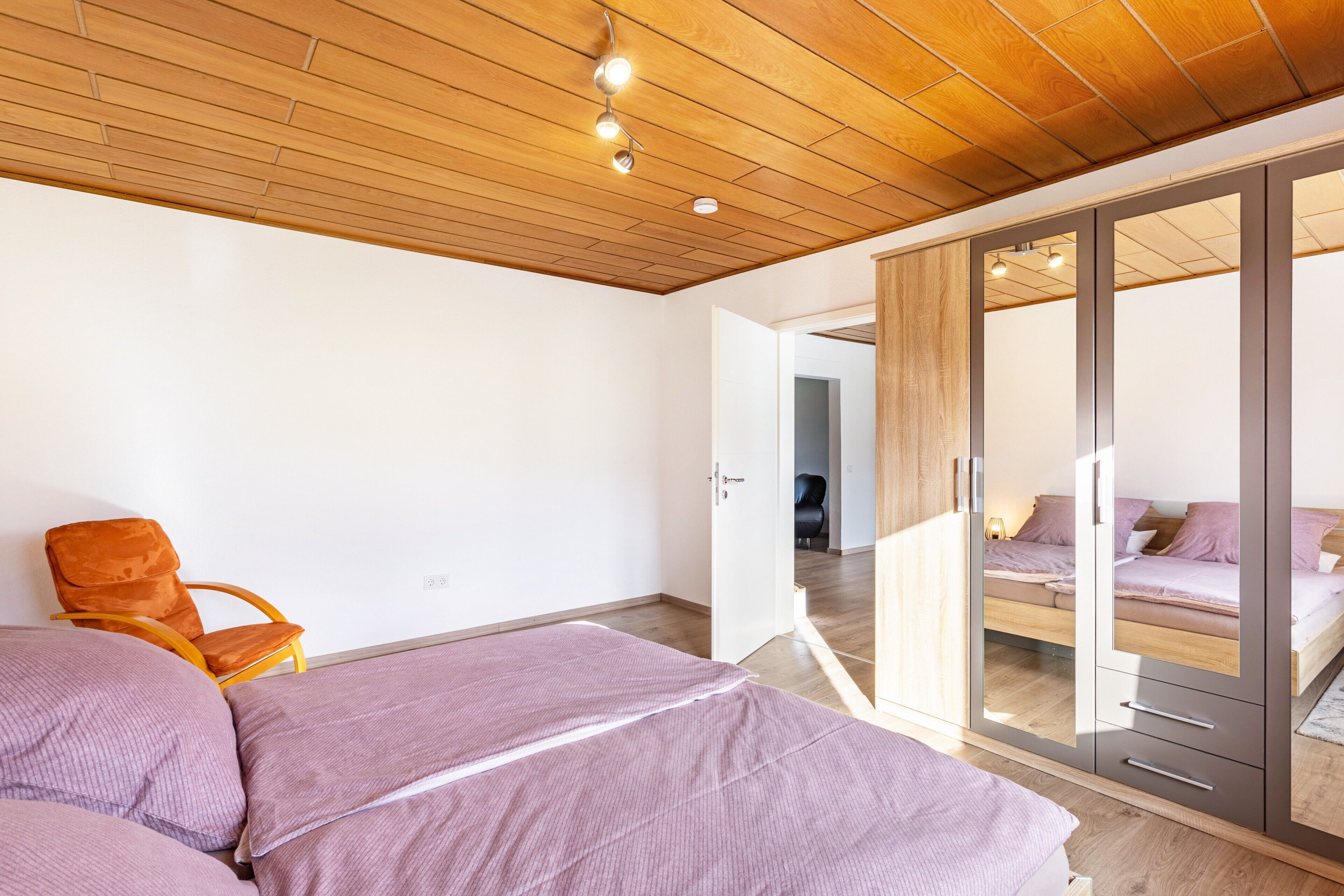 Un dormitorio con techo de madera, una cama con ropa de cama rosa, una silla y un armario con puertas de espejo.