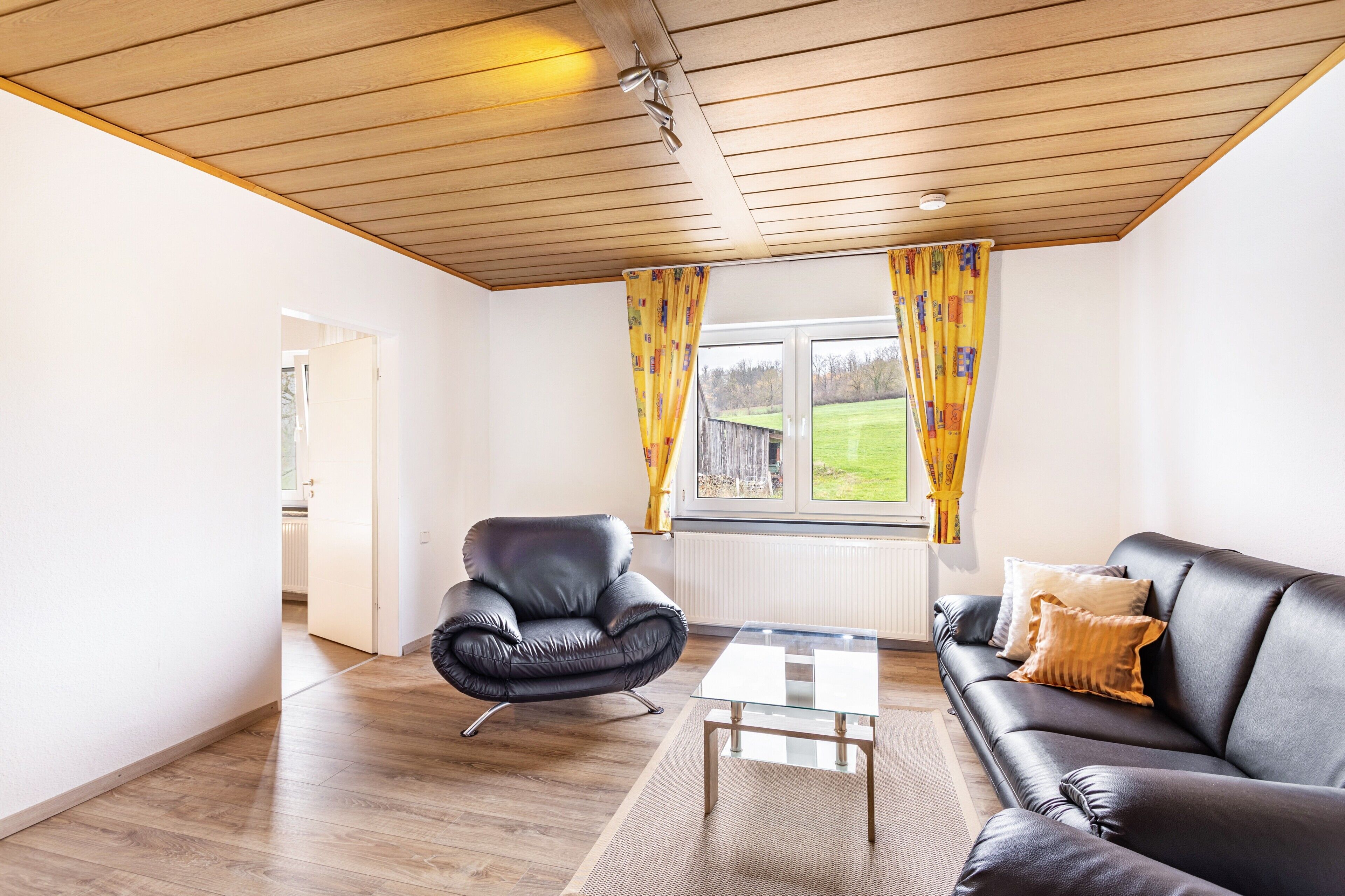 Un salon avec un plafond en bois, un canapé en cuir noir, une table basse en verre, un fauteuil en cuir noir et une fenêtre avec des rideaux jaunes.