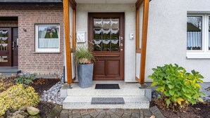 Ett hus med en fasad av tegel, en trädörr med glaspaneler, en krukväxt och en liten trädgårdsyta med stenar och växter.