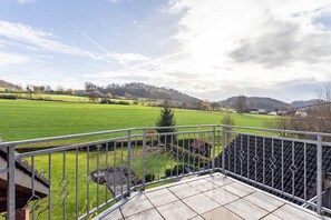Un balcon doté d’une rambarde en métal, donnant sur un champ verdoyant et une colline lointaine.