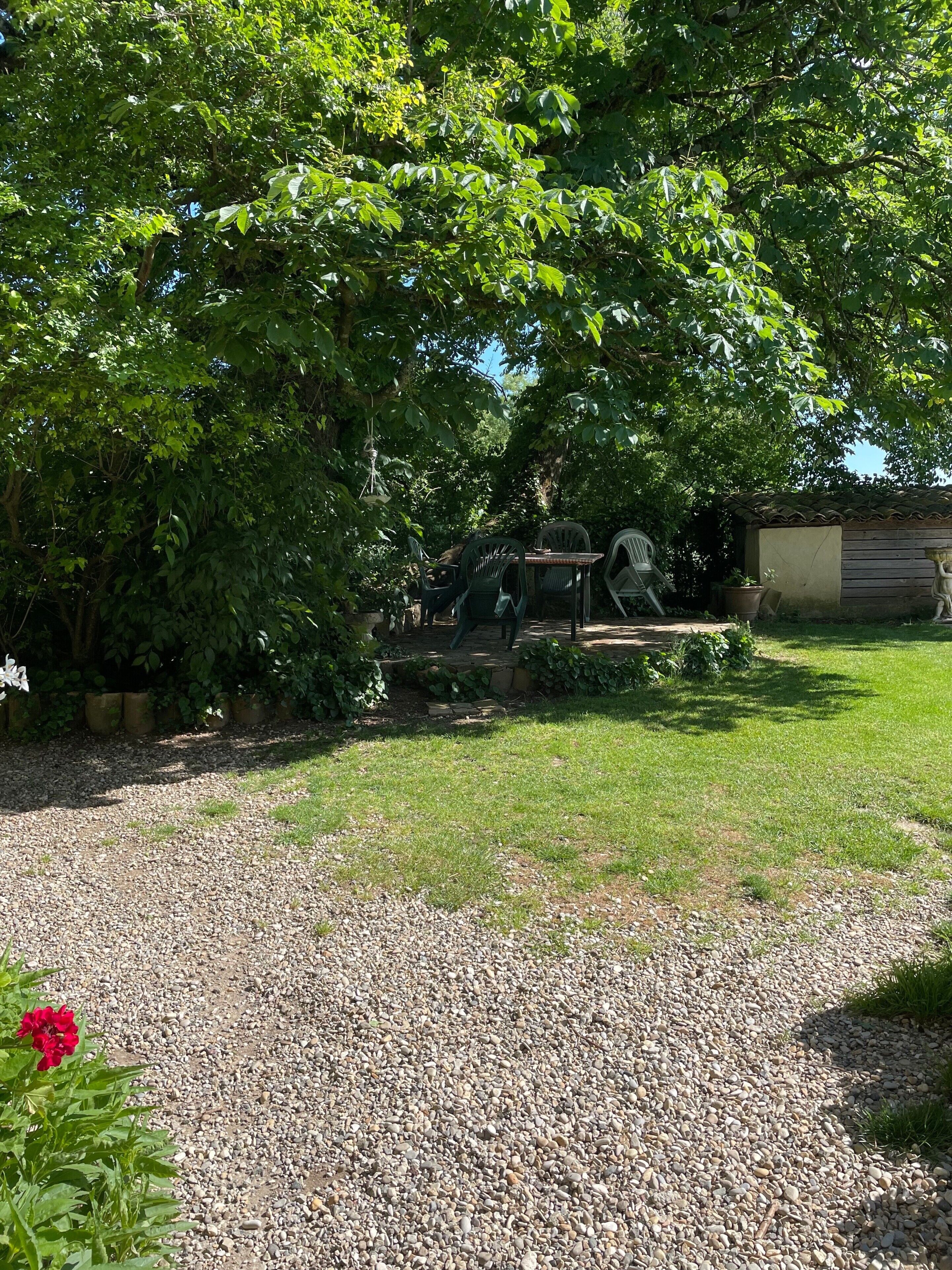 A gravel path leading to a patio area with outdoor furniture, surrounded by lush greenery and a small building with a tiled roof.