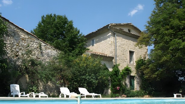 Une piscine entourée de chaises longues, un bâtiment en pierre et une végétation luxuriante.