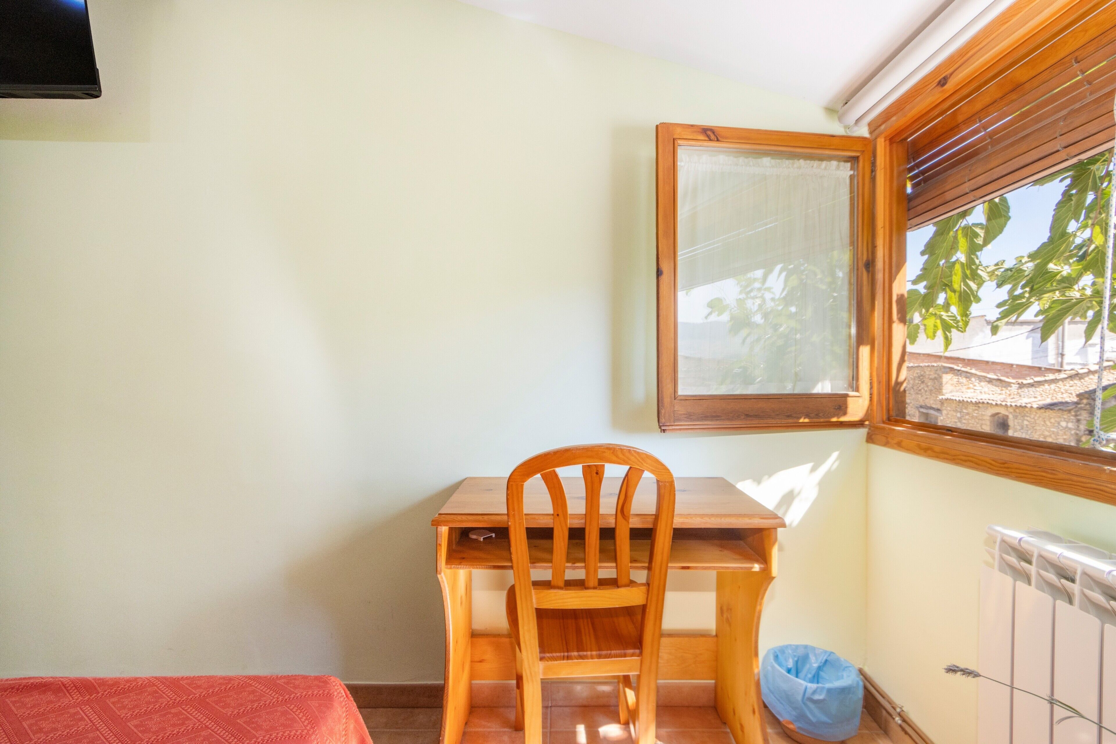 Una habitación pequeña con un escritorio y una silla de madera, una ventana con persianas, un radiador y una bolsa azul en el suelo.