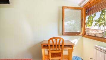 Una habitación pequeña con un escritorio y una silla de madera, una ventana con persianas, un radiador y una bolsa azul en el suelo.