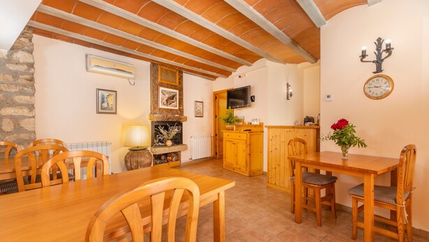 A dining area with wooden tables and chairs, a kitchen with wooden cabinets, a stone wall, and a clock on the wall. - Room 'Habitació Triple' with Mountain View, Wi-Fi and Air Conditioning (Conques)