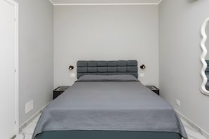 Een slaapkamer met een grijs bed, een gestoffeerd hoofdeinde, nachtkastjes en een spiegel aan de muur.
