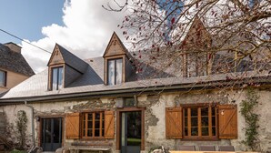 A stone building with a sloped roof, wooden shutters, and a dining area with chairs.