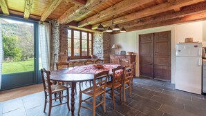 A rustic kitchen with a stone wall, wooden ceiling beams, a dining table with chairs, and a refrigerator.