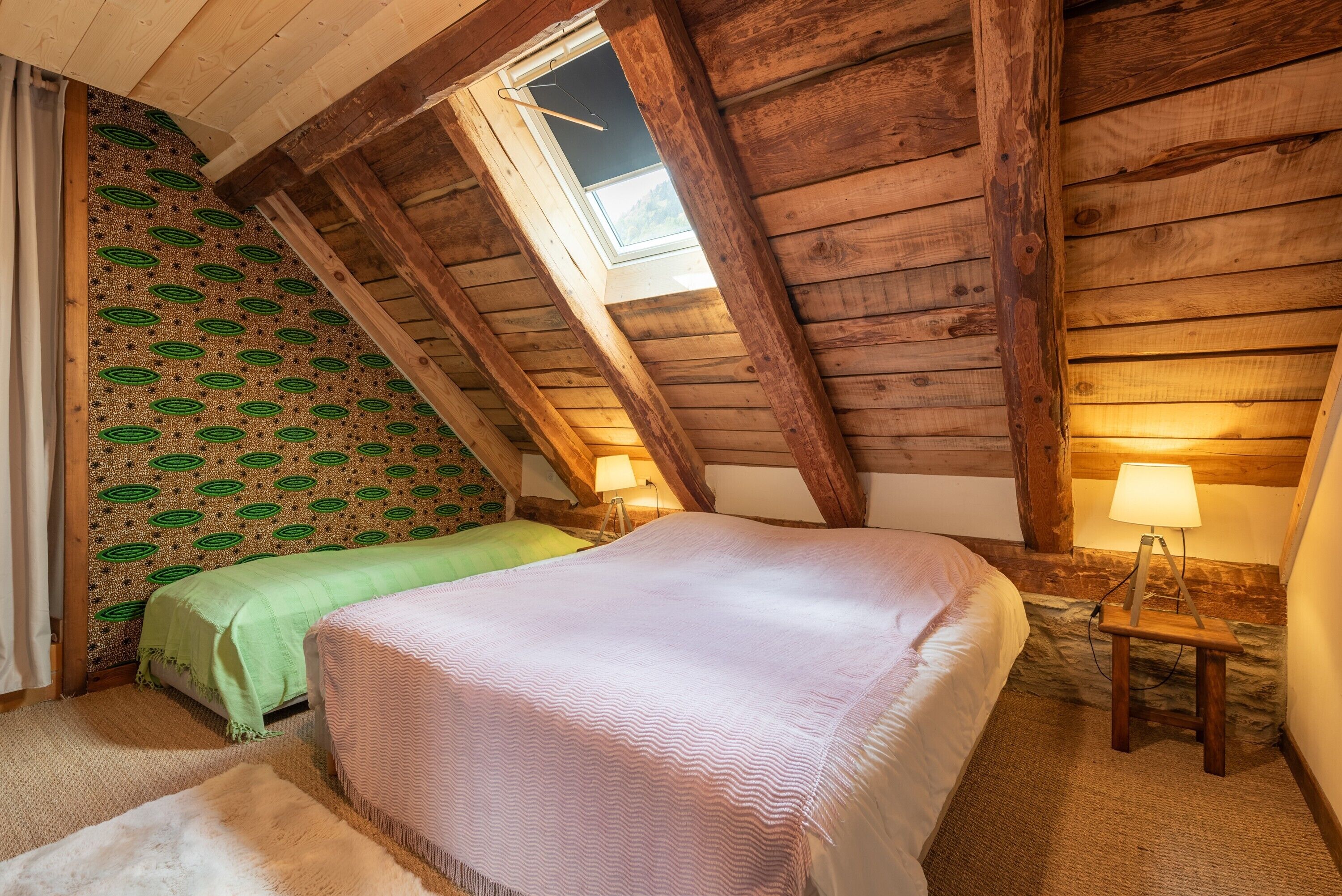 Une chambre mansardée chaleureuse avec un plafond en pente, deux lits, un puits de lumière et une petite table avec une lampe.