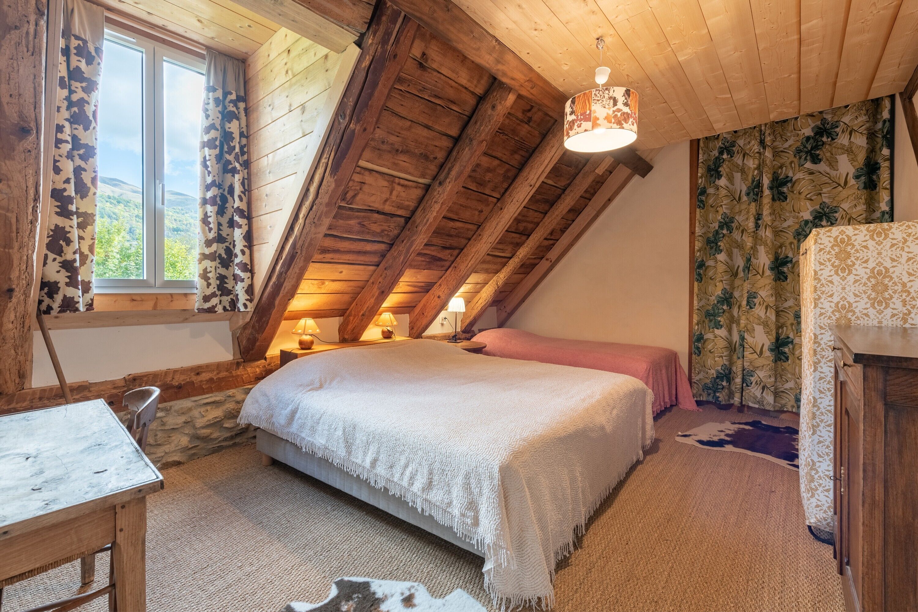 Ein Schlafzimmer mit schrägen Wänden, einem Bett mit weißer Bettdecke, einem Holztisch und einem Fenster mit Vorhängen.