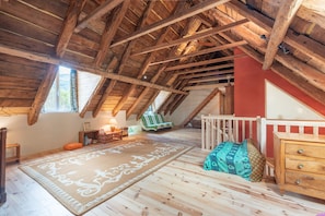 A loft bedroom with exposed wooden beams, a sloped ceiling, a large window, a wooden dresser, a bed with patterned bedding, and a decorative rug.