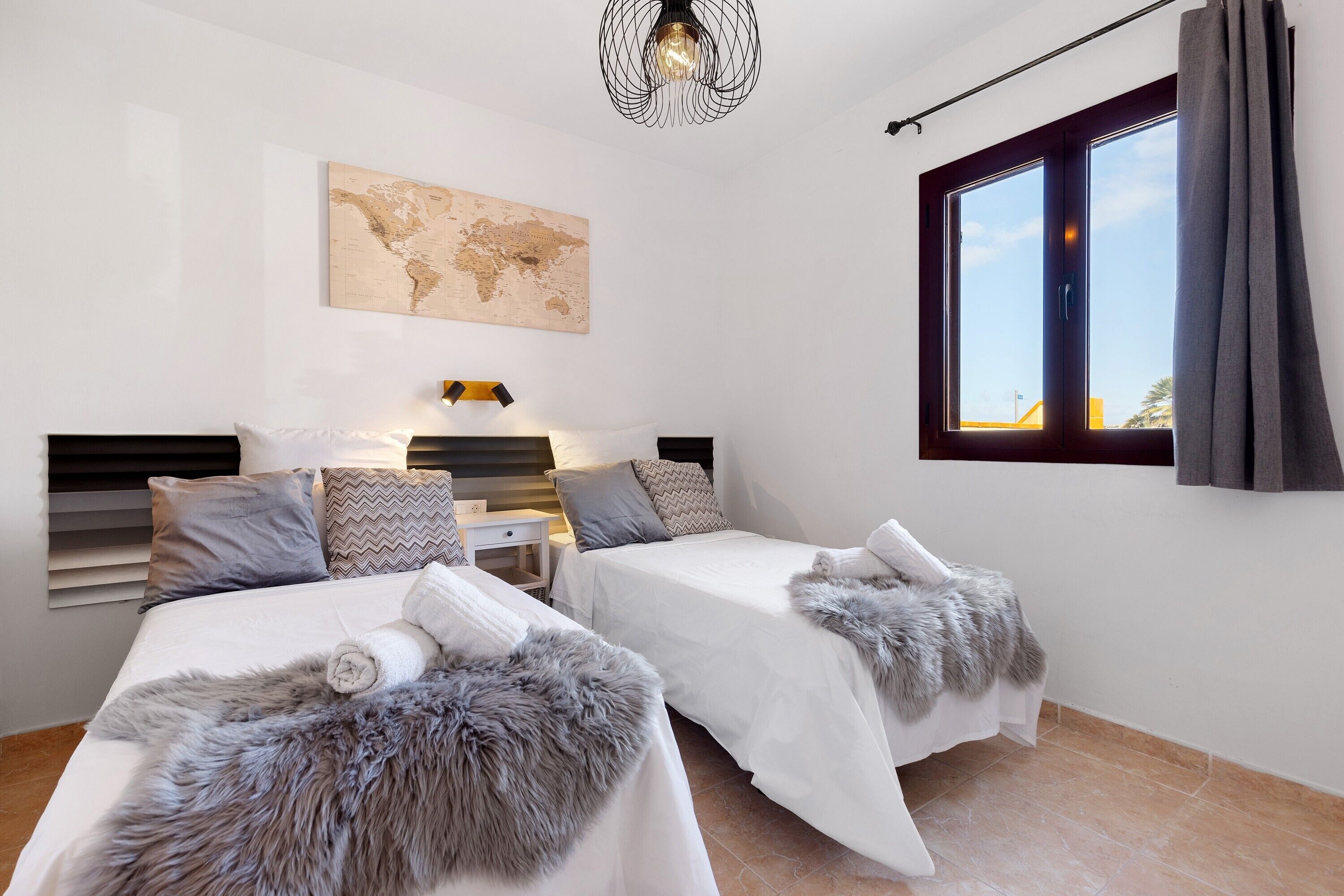 A bedroom with two beds, a world map on the wall, a window with curtains, and a hanging light fixture.