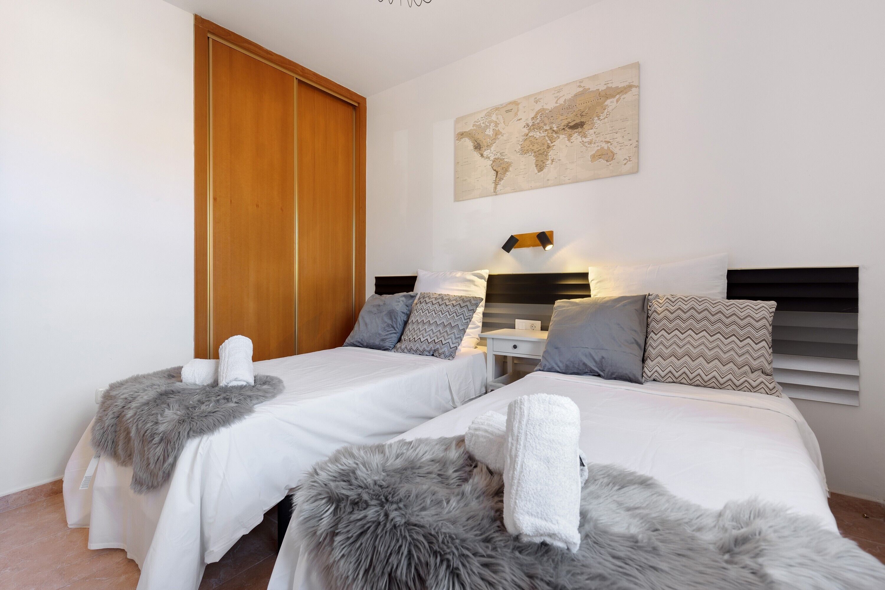 A hotel room with two beds, a wooden wardrobe, a world map on the wall, and a nightstand with a lamp.