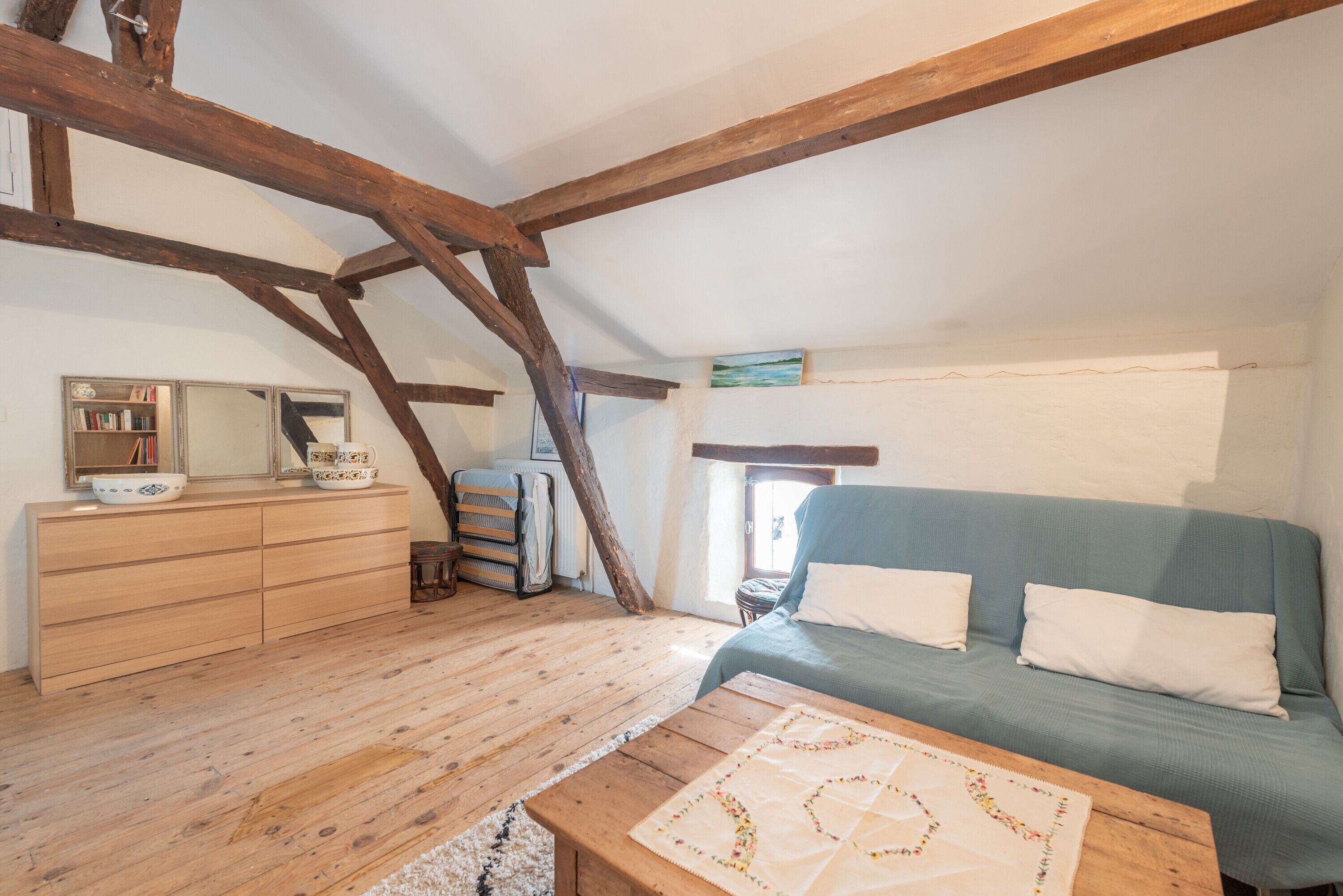 A cozy attic bedroom with exposed wooden beams, a wooden dresser, a blue sofa, and a quilt on the table.