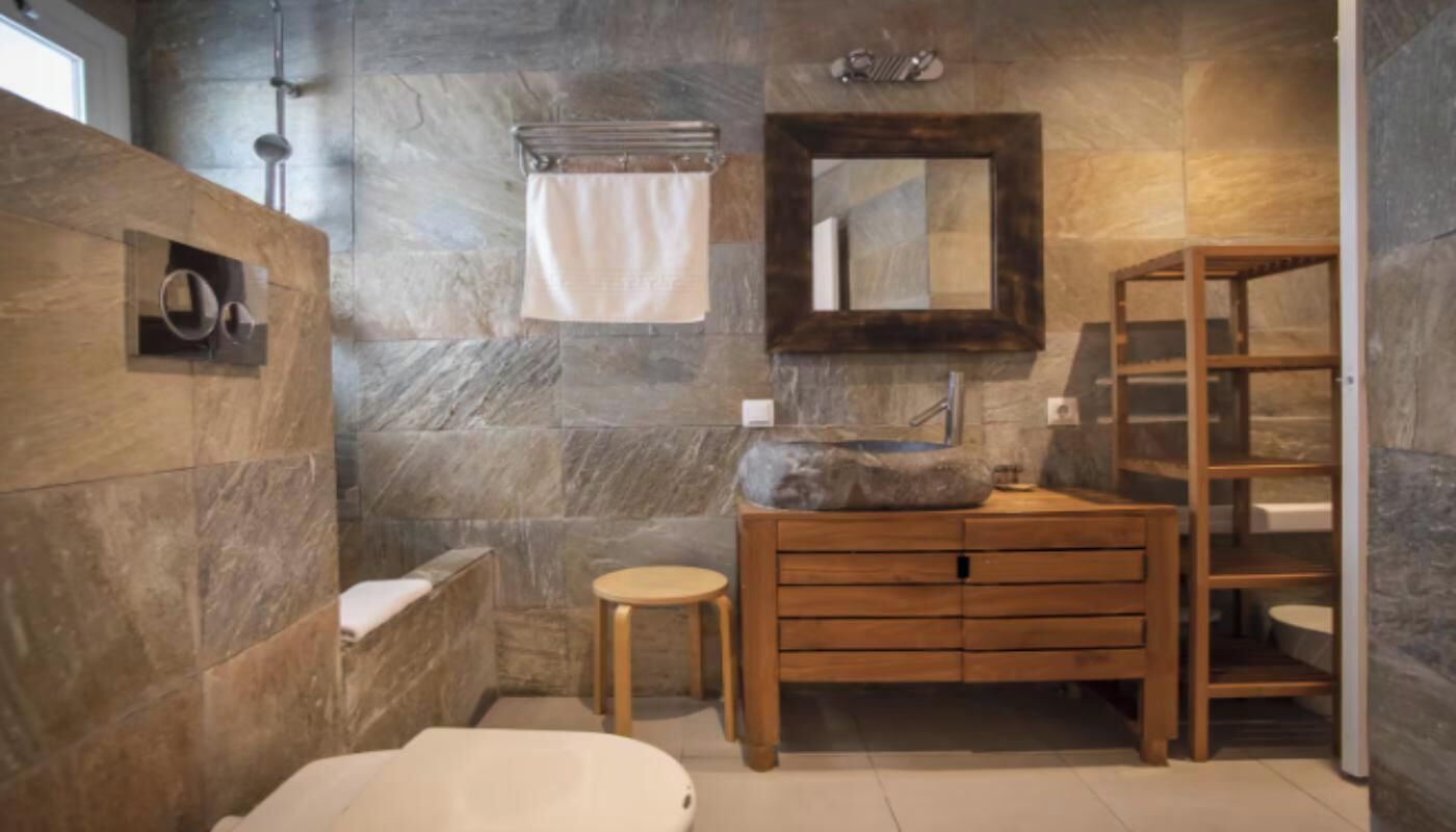 A bathroom with a stone wall, wooden vanity, a round stool, and a mirror.