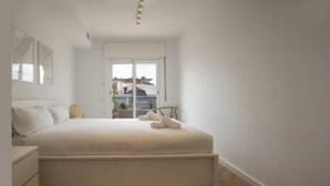 A bedroom with a bed, a nightstand, a bedside table, and a window with a view of buildings. - Spectacular Penthouse with Sagrada Familia Views (Barcelona)
