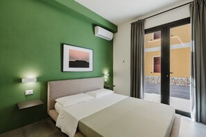 Et hotelværelse med en seng, en fastgjort aircondition, et indrammet billede, et natbord med en lampe og udsigt gennem en glasdør.
