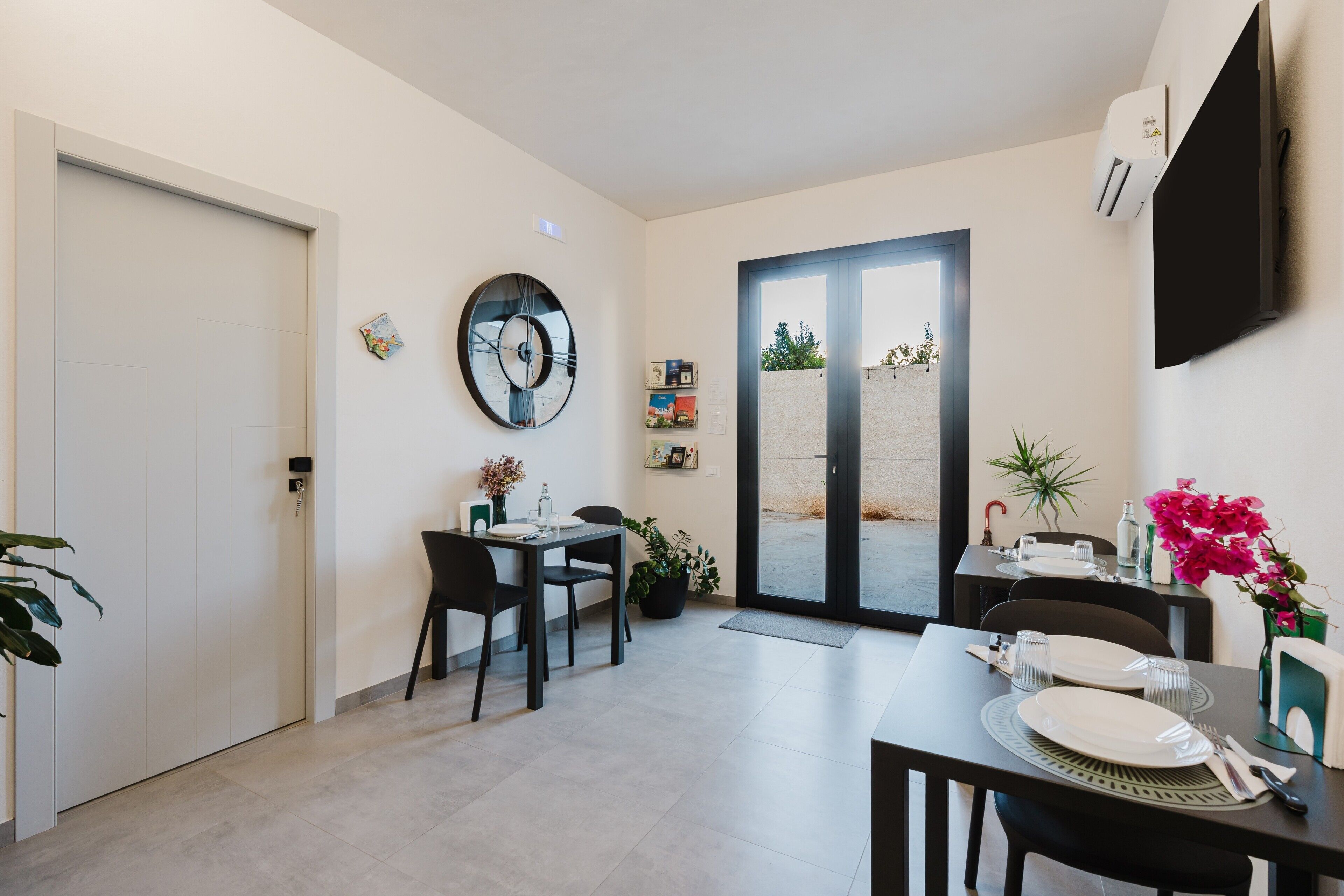 Un comedor moderno con una mesa dispuesta para dos personas, un reloj en la pared y un ventanal corredero que da al exterior.