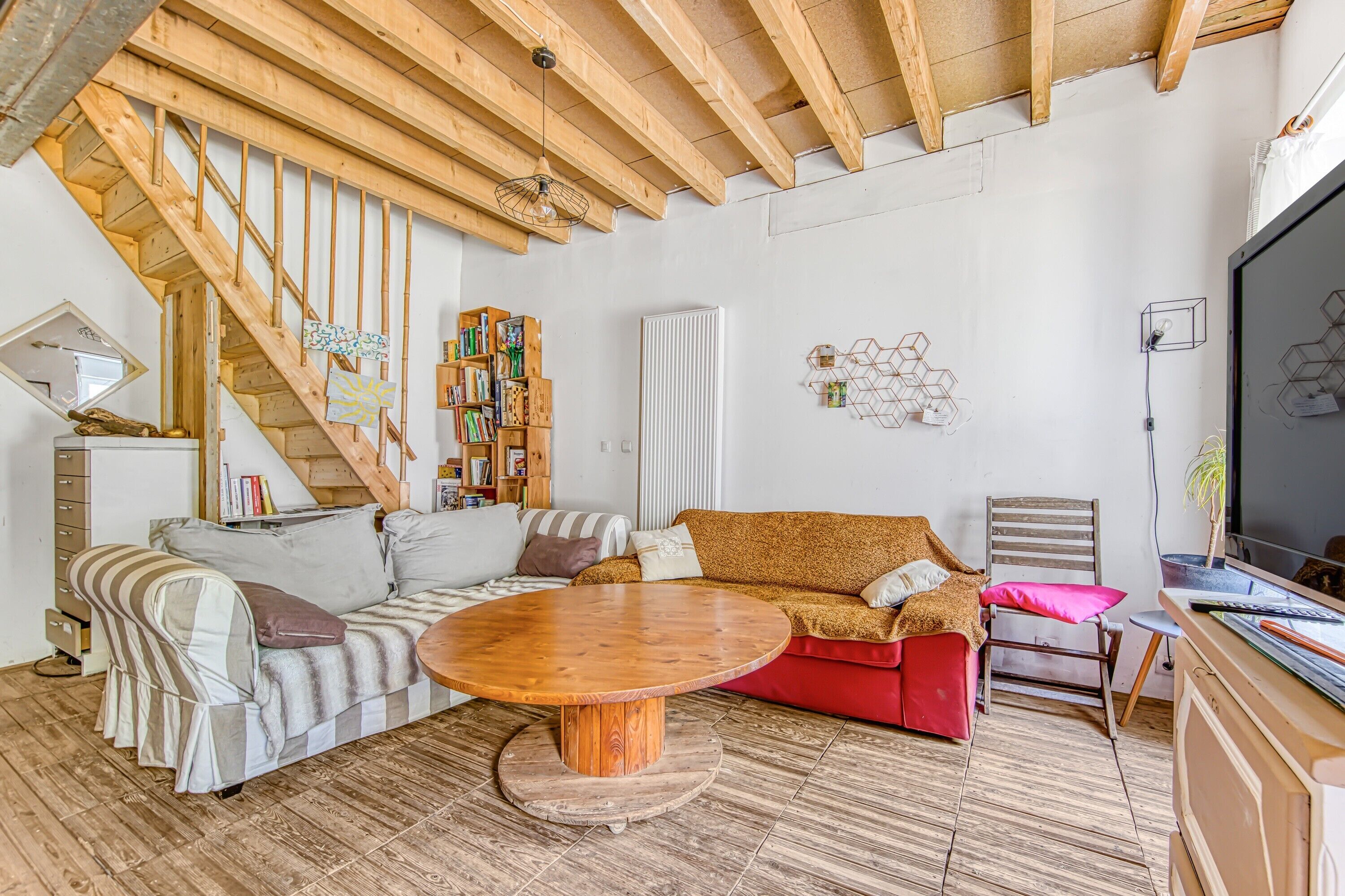 Un salon avec un escalier en bois, une table basse ronde en bois, un canapé, un fauteuil rouge et une bibliothèque.