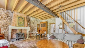 A rustic interior with exposed wooden beams, a stone wall, a fireplace, a round wooden table, and a staircase.