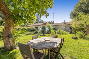 Una mesa y sillas de madera en un jardín con un árbol, un muro de piedra y una casa al fondo.