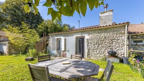 A stone building with a tiled roof, a wooden table with chairs, and a garden area.