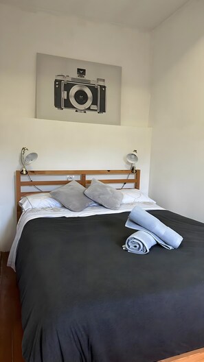 Una cama con una colcha oscura, dos almohadas, una toalla enrollada y una cámara en la pared de arriba.