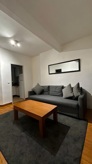 Una sala de estar con un sofá gris, una mesa de centro de madera y una cocina visible a través de un vano abierto.