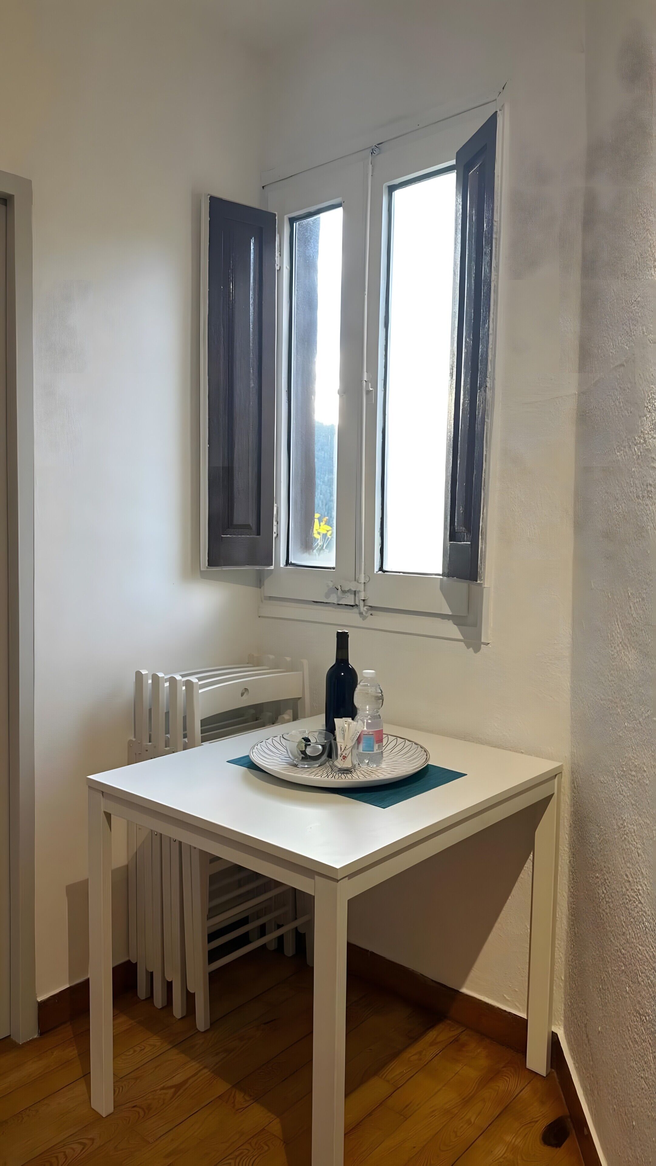 Een kleine kamer met een witte tafel, een raam met blauwe luiken, een radiator en een houten vloer.
