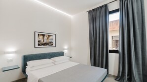 Quarto de hotel moderno com cama, mesa de cabeceira, luminária na parede, quadro emoldurado e janela com cortinas.