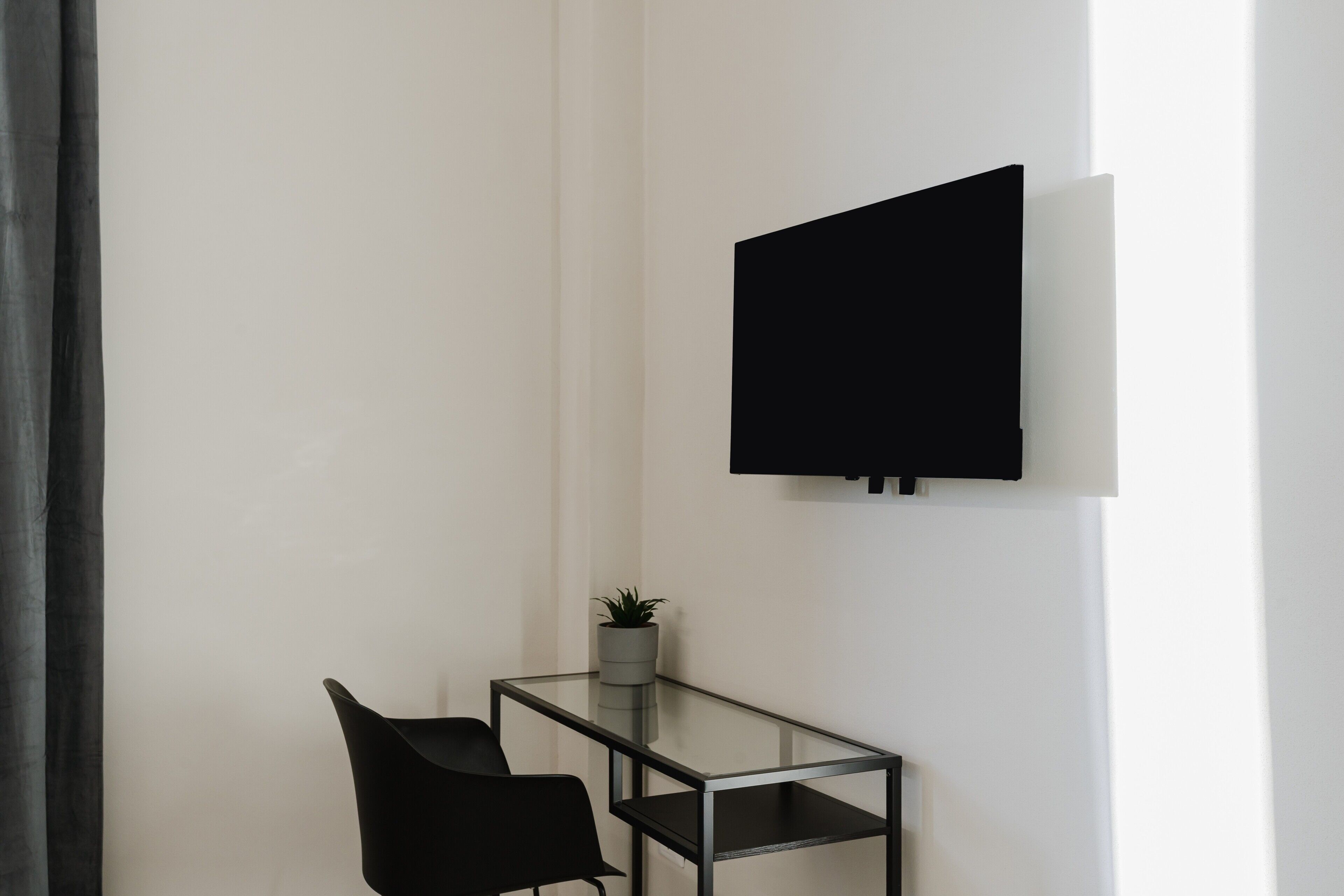 Una habitación moderna con una mesa de vidrio, una silla negra, una planta en maceta y un televisor montado.