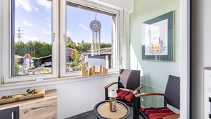 A room with two large windows, a wicker chair with a cushion, a small table with a bottle and a candle, and a framed picture on the wall.