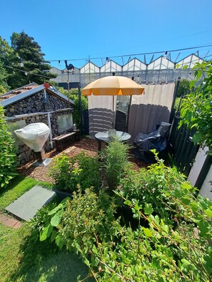 Un jardín con una mesa y sillas bajo una pérgola, rodeado de plantas y un muro de piedra.